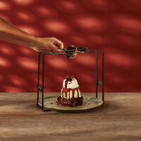 Brownie de chocolate com sorvete e chantilly e raspas de chocolate, com uma mão servindo calda de chocolate