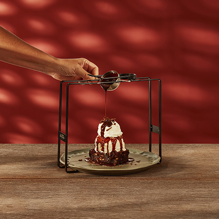 Brownie de chocolate com sorvete e chantilly e raspas de chocolate, com uma mão servindo calda de chocolate