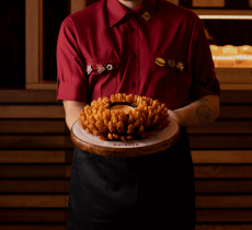 Um homem com um uniforme vermelho, segurando um prato com as batatas do Outback, no centro do prato tem um molho da casa.