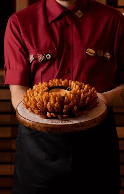 Um homem com um uniforme vermelho, segurando um prato com as batatas do Outback, no centro do prato tem um molho da casa.