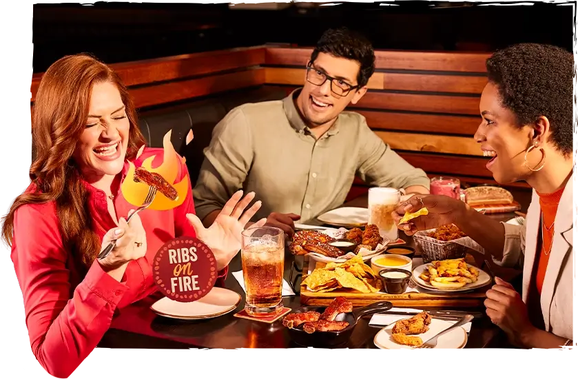 Amigos participando da promoção Robs on Fire, mulher segura costela flamejante enquanto homem e mulher observam sorrindo.