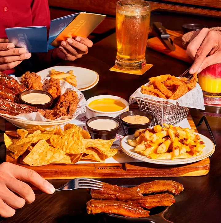 Pessoas compartilhando uma variedade de petiscos no Outback, incluindo wings, costelas, batatas e nachos, com bebidas como cerveja e coquetel.