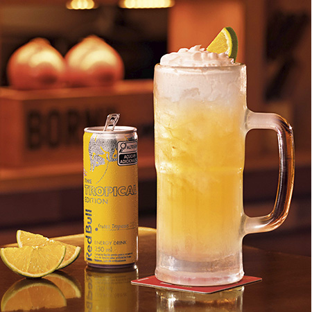 Caneca de drink não alcoólico Tropical Summer, ao lado de uma lata de Red Bull Tropical e fatias de limão.